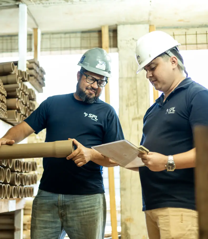 spl slide 2 Dois colaboradores da SPL utilizando capacetes de segurança inspecionam um tubete de papelão em um estoque enquanto um deles confere anotações em uma prancheta.